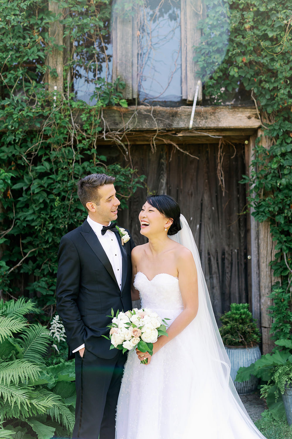 A groom and laughing bride holding a white bridal bouquet by Bedford Village Flower Shoppe